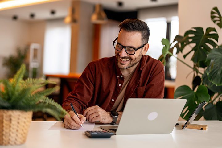 bookkeeping A person wearing glasses and a red shirt is smiling while writing at a desk with a laptop, tablet, and calculator. A plant is nearby.