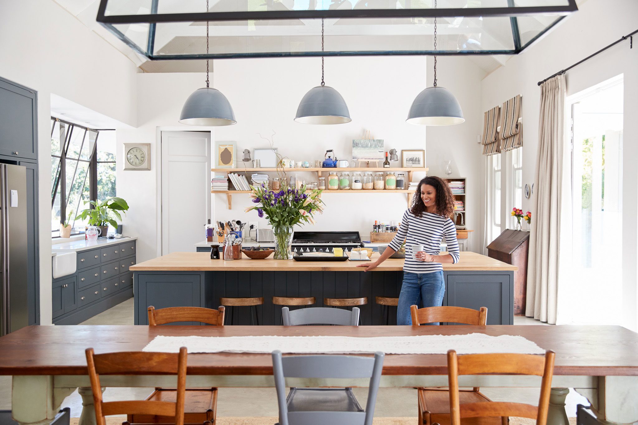 shutterstock_1065427163 A woman stands in a bright, modern kitchen holding a mug, near an island with flowers and shelves filled with jars and kitchenware.