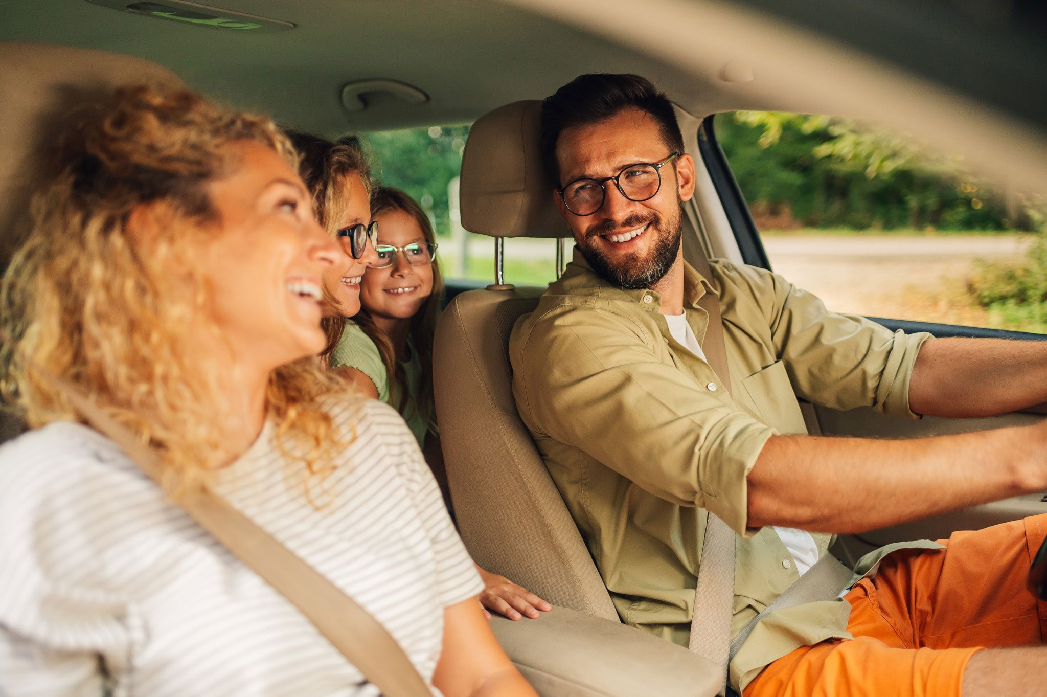 shutterstock_2415342603 A man drives a car with a woman in the front passenger seat and two children in the back, all smiling and wearing seatbelts.