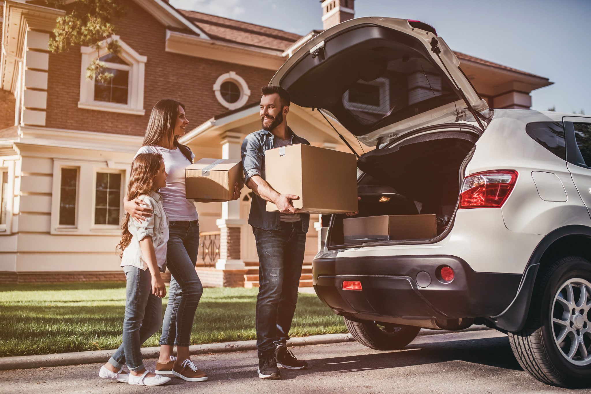 shutterstock_669633361 A family stands by a car with its trunk open, holding cardboard boxes in front of a suburban house, suggesting moving or unpacking.